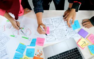desk filled with papers, colourful post-its, and showing several hands reaching in holding pens and writing
